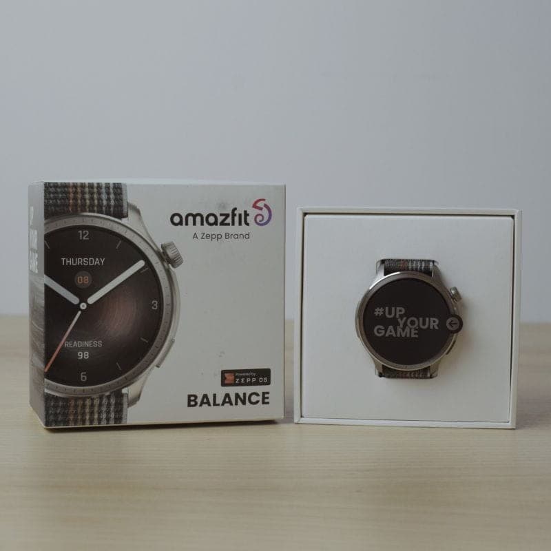 Amazfit Balance cinzento com caixa prateada, mostrador redondo 1,5 polegadas AMOLED e bracelete têxtil listrada; embalagem ao lado do relógio exibido