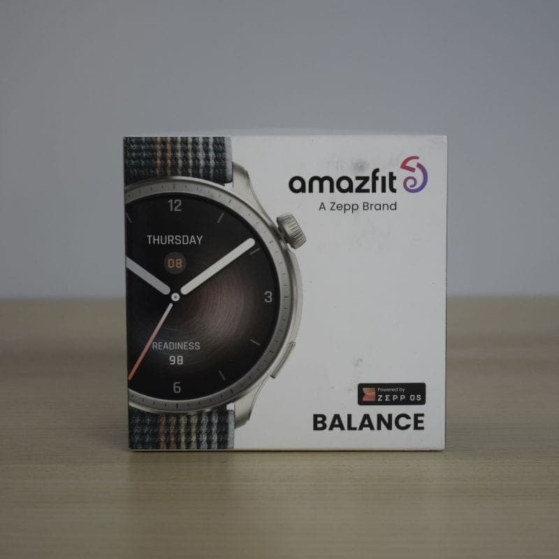 Caixa do Amazfit Balance cinzento com mostrador redondo 1,5 AMOLED, moldura metálica escovada e bracelete têxtil multicolor sobre fundo neutro
