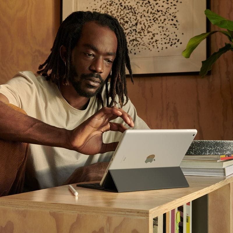 iPad 10,2 argent sur étui support, écran allumé, Apple Pencil posé à côté, ambiance intérieure chaleureuse avec déco murale et bois