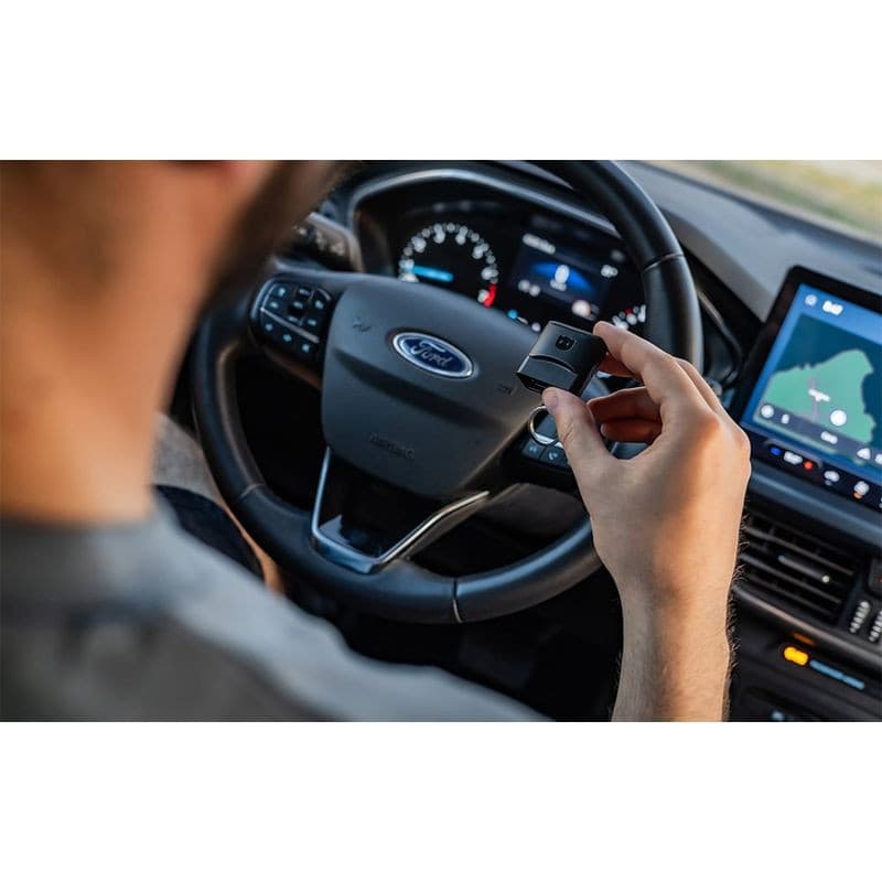 Scanner de diagnostic OBDeleven 3 Ultimate Pack tenu en main dans une voiture Ford, petit dongle noir OBD2, tableau de bord et écran GPS visibles.