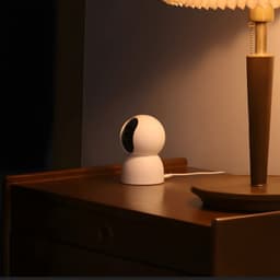 Xiaomi Smart Camera C500 6MP 3,5K WiFi 6 posée sur table de chevet, design blanc arrondi, lentille noire orientable, câble branché, lumière chaude ambiante