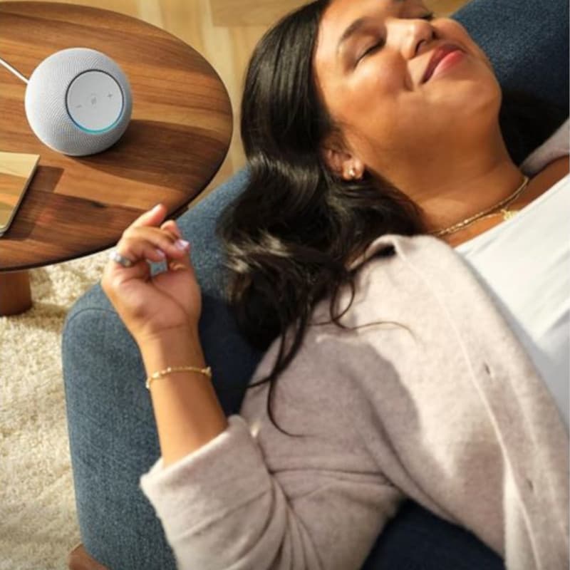 Amazon Echo Dot Max 2025 Grafite em mesa de madeira, corpo arredondado cinza, topo com LEDs azuis e botões, design compacto ao lado de sofá