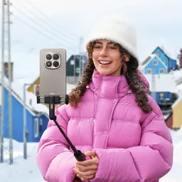 Jovem com casaco rosa usa bastão de selfie com smartphone Xiaomi Redmi Note 15 Pro 5G dourado, quatro câmeras traseiras grandes, cenário de neve ao fundo