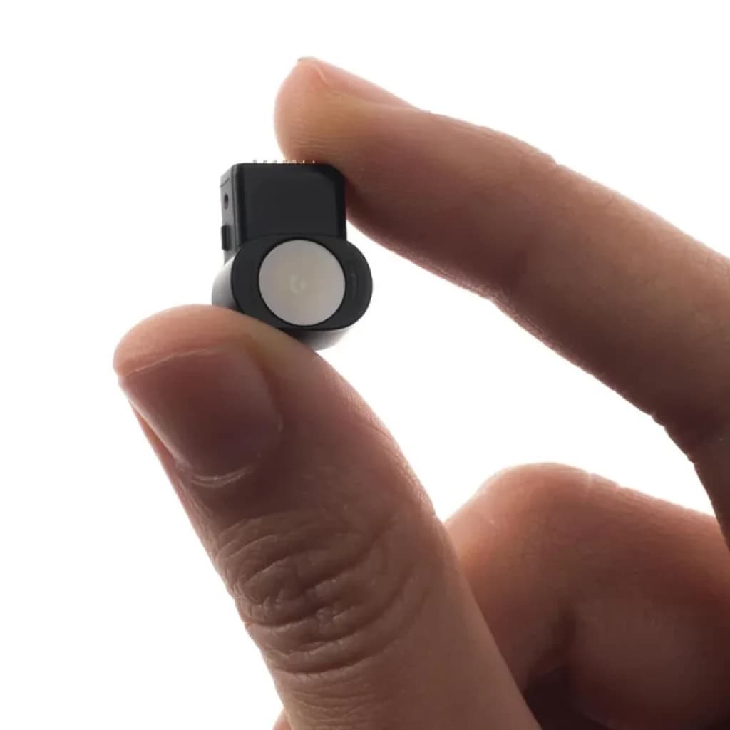 Module caméra XREAL Eye noir avec lentille circulaire blanche, tenu entre les doigts pour montrer sa petite taille et son design minimaliste.
