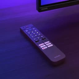Télécommande Philips élégante posée sur table sombre, éclairage ambilight violet bleu, touches Netflix Prime Video YouTube et pavé numérique bien