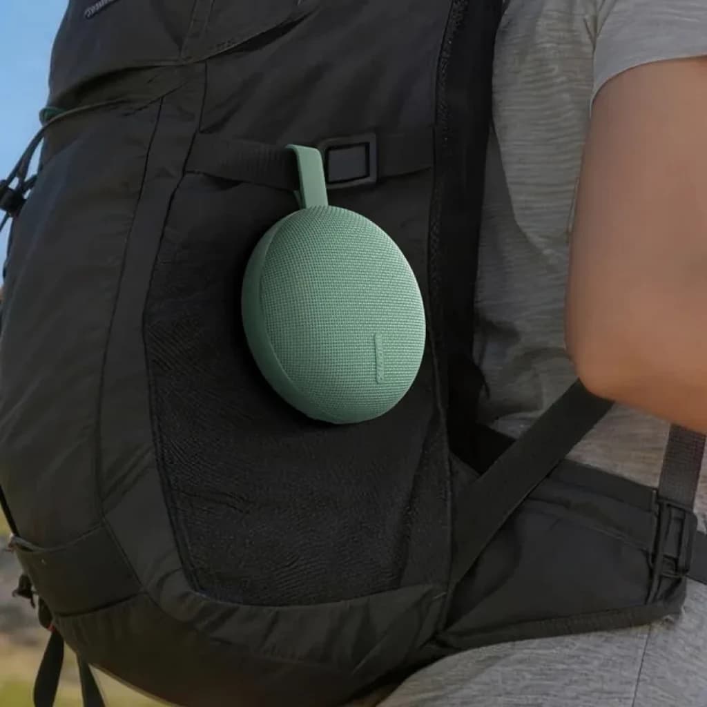 Enceinte portable ronde vert menthe accrochée à un sac à dos noir, tissu maillé, sangle de transport, compacte et légère pour usage extérieur
