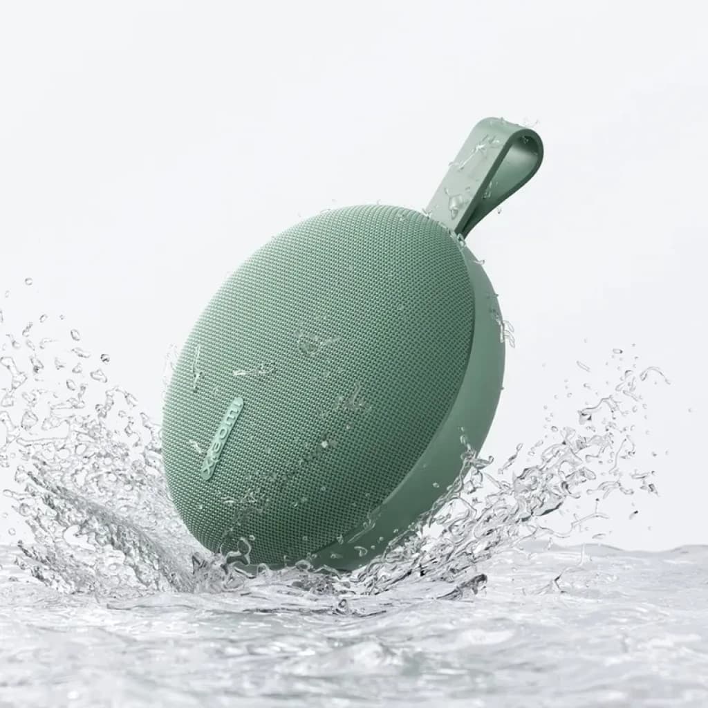 Enceinte portable ronde vert sauge avec sangle, grille fine, résistante à l’eau IP66, format compact, éclaboussures visibles, design minimal pour écoute