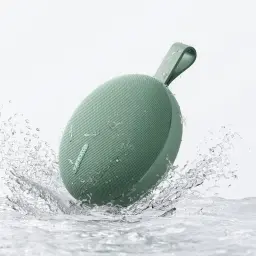 Enceinte portable ronde vert sauge avec sangle, grille fine, résistante à l’eau IP66, format compact, éclaboussures visibles, design minimal pour écoute