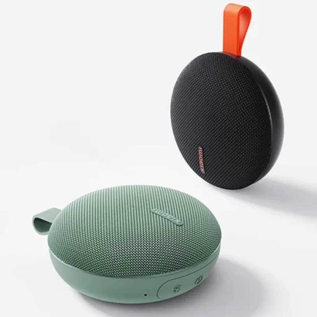 Enceinte portable ronde vert menthe avec maille fine, logo Xiaomi, boutons latéraux et languette d’accroche. Modèle noir au fond avec sangle orange. IP66.