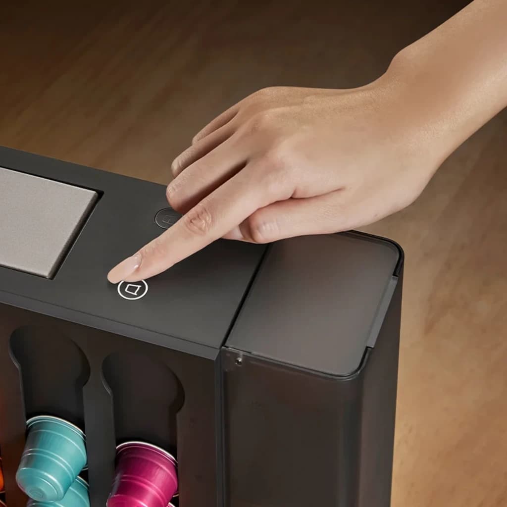 Main appuyant le bouton tactile sur le dessus noir; compartiment latéral translucide et rangement vertical de capsules colorées visibles en façade