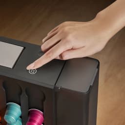 Main appuyant le bouton tactile sur le dessus noir; compartiment latéral translucide et rangement vertical de capsules colorées visibles en façade