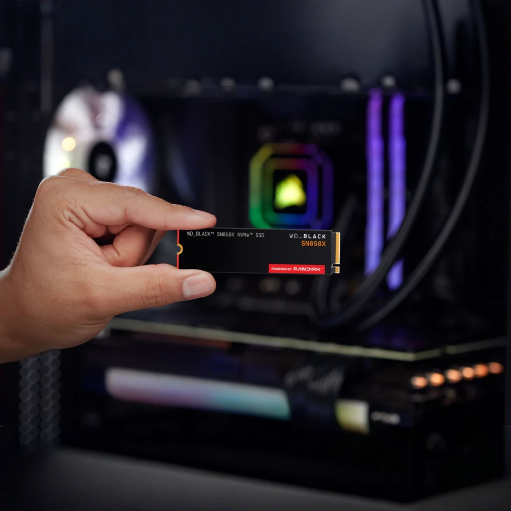 SSD WD_BLACK SN850X 2 To NVMe PCIe 4.0 M.2 2280, en situation: main tenant le SSD devant un PC gaming éclairé RGB