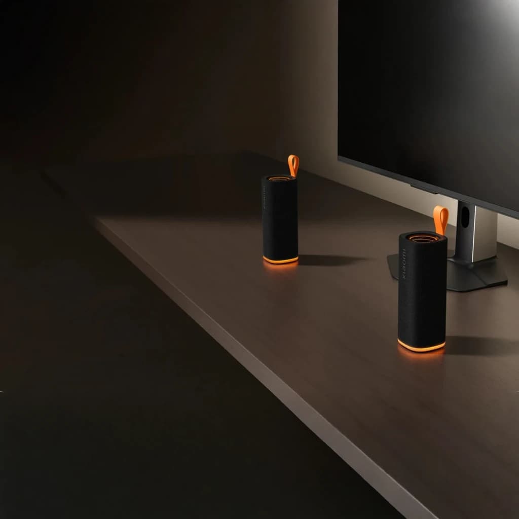 Altavoz Bluetooth Xiaomi Sound Play negro cilíndrico con malla textil, base y asa naranja, luz LED inferior, diseño compacto junto a monitor en escritorio