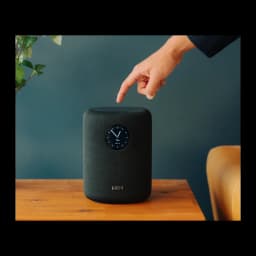 Wiim Sound negro, uso: mano señalando la parte superior, pantalla circular con reloj visible en tela negra sobre mesa de