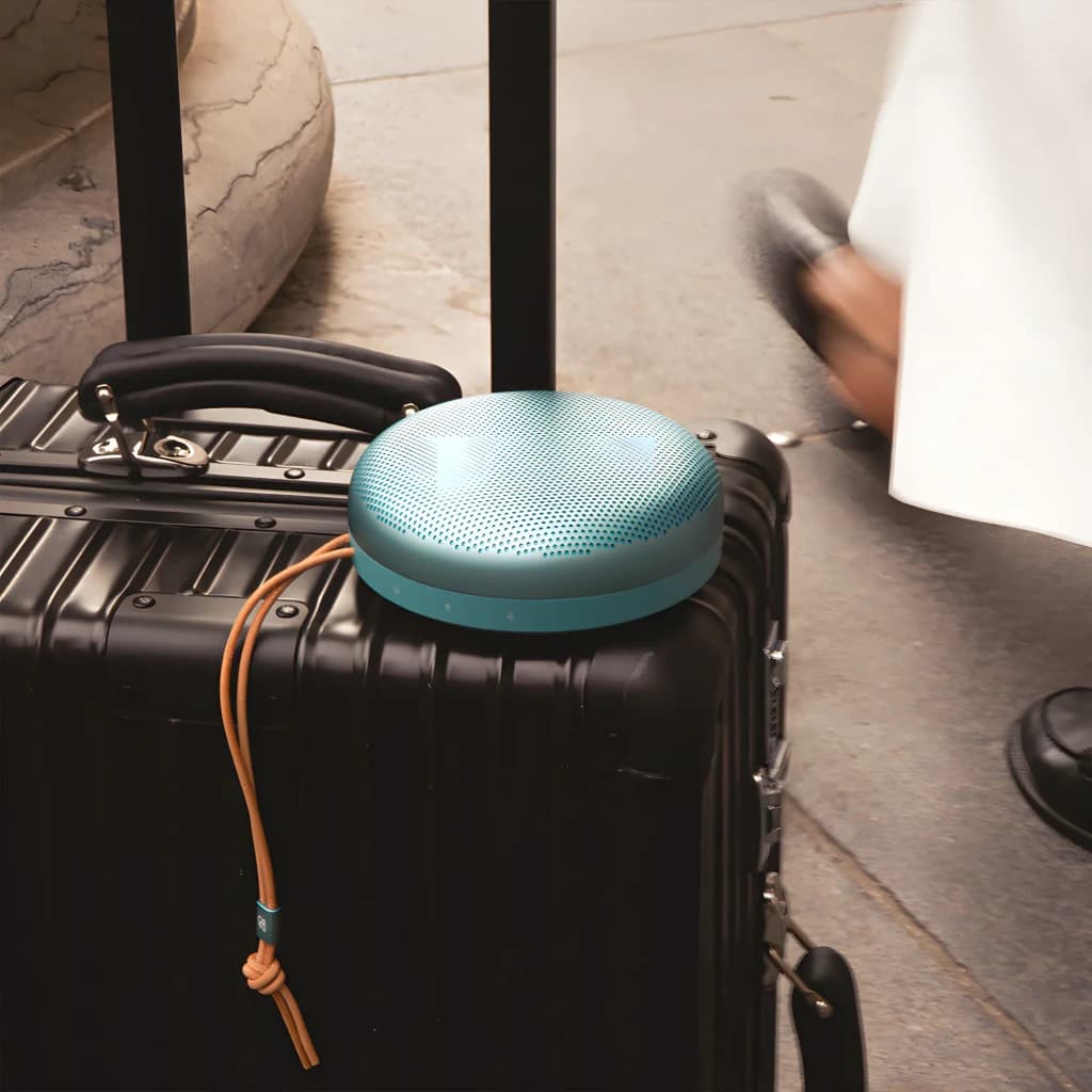 Bang & Olufsen Beosound A1 3ª Gen verde eucalipto, vista lifestyle: altavoz sobre maleta negra con correa de cuero en