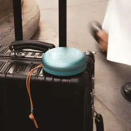 Bang & Olufsen Beosound A1 3ª Gen verde eucalipto, vista lifestyle: altavoz sobre maleta negra con correa de cuero en