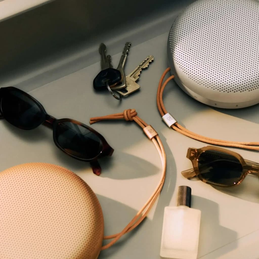 Bang & Olufsen BeoSound A1 3rd Gen Rojo Rústico, lifestyle: dos altavoces con correa de cuero junto a gafas y llaves