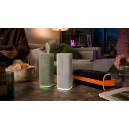 Altavoz portátil verde con forma rectangular, rejilla textil clara, base luminosa, asa de silicona y diseño resistente, junto a variante en gris y funda