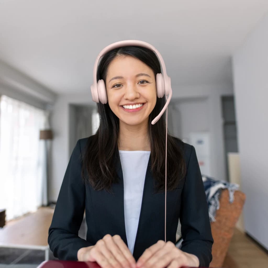 Logitech Auriculares H390 USB Rosa, uso: mulher sorridente com headset rosa e microfone em ambiente de trabalho remoto
