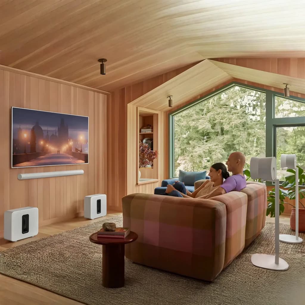 Sonos Sub Gen 4 branco, cena sala de estar: subwoofer branco junto ao sofá com casal a ver TV, sistema home cinema completo