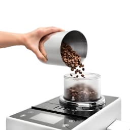 De'Longhi Rivelia EXAM440.35B Branco, detalhe tolva: mão despejando grãos de café no reservatório transparente sobre painel