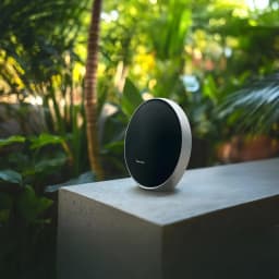 Harman Kardon Onyx Studio 9 negro, vista frontal: altavoz oval sobre superficie de hormigón con fondo de vegetación tropical