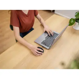 Trust Voxx Ratón Ergonómico Negro, uso en escritorio: mujer sujetando el ratón junto a portátil plateado sobre mesa de madera