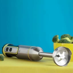 Jata Liquidificadora JEBT1789 Inox, vista lateral: varinha deitada em superfície amarela com lâmina visível e tigela de