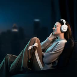SteelSeries Arctis Nova Pro Omni branco, uso: mulher relaxada em cadeira com auriculares, fundo urbano noturno