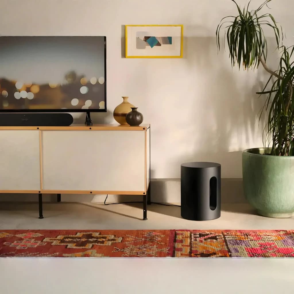 Sonos Sub Mini noir, vue salon: caisson cylindrique près d'un meuble TV avec barre de son et plante