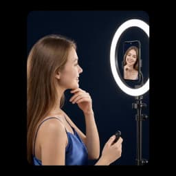 Anneau lumineux LED circulaire sur trépied, smartphone au centre affichant un selfie, télécommande en main, fond bleu nuit, éclairage doux et homogène
