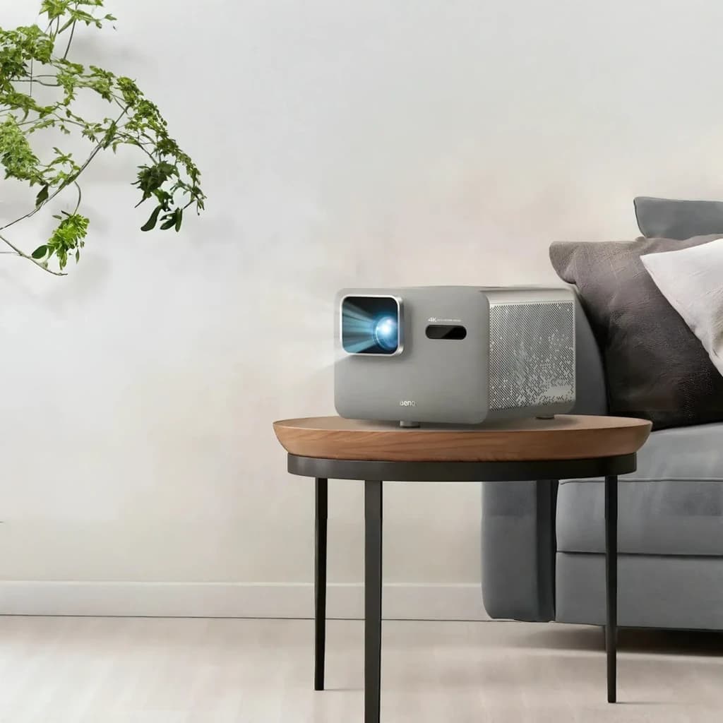 BenQ Proyector TK705STi 4K UHD 3000lm Gris, vista frontal en salón: lente iluminada sobre mesa auxiliar junto a sofá