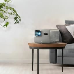 BenQ Proyector TK705STi 4K UHD 3000lm Gris, vista frontal en salón: lente iluminada sobre mesa auxiliar junto a sofá