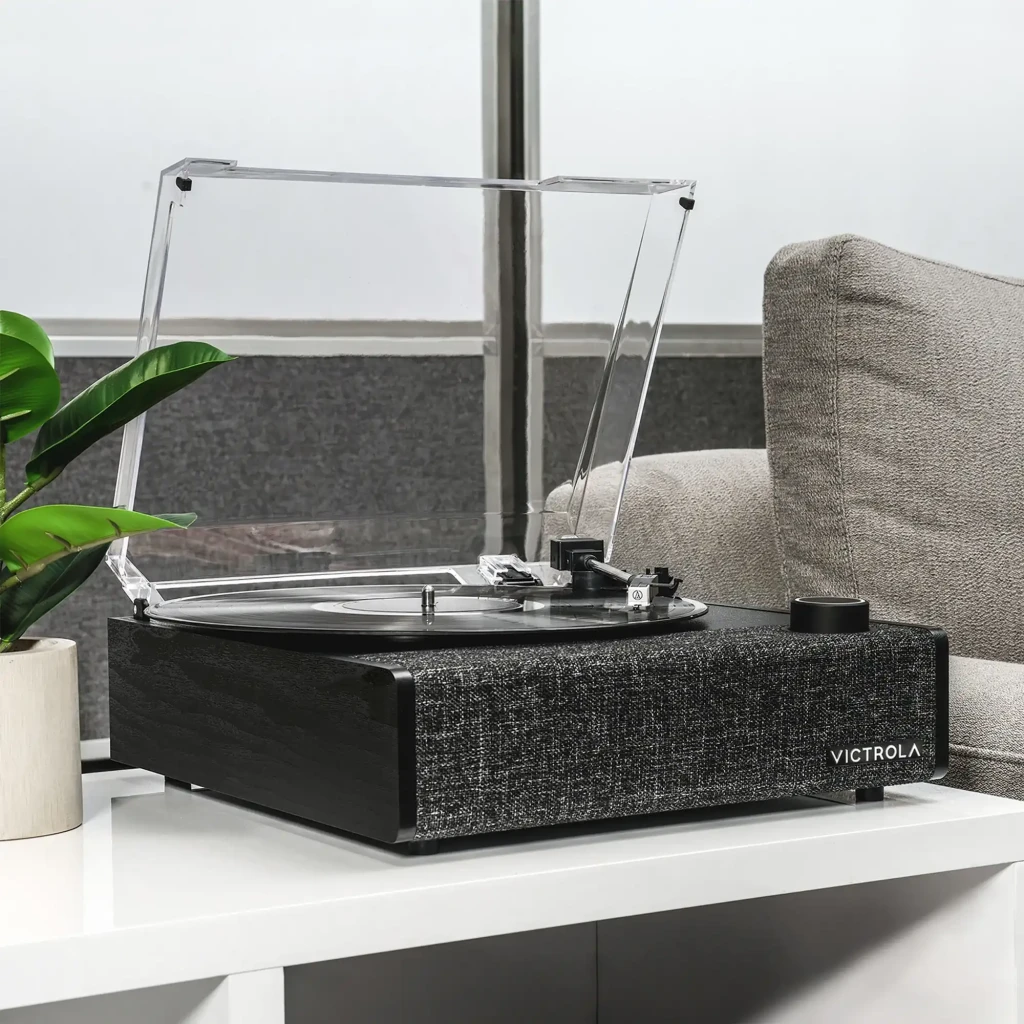 Tocadiscos Victrola Eastwood II VTA-74 con tapa transparente, plato negro, brazo recto y base textil gris; perilla frontal y logotipo Victrola visibles - Ítem3