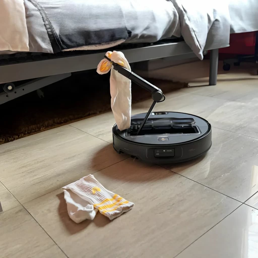 Roborock Saros Z70 negro, en uso: brazo robótico recogiendo un calcetín del suelo junto a una cama