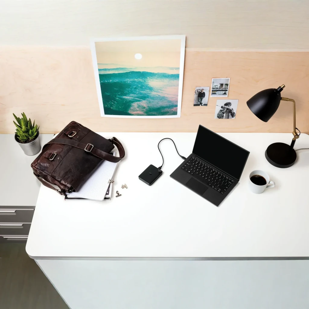 WD Elements SE WDBJRT0050BBK 5TB preto, uso: conectado por USB a laptop sobre mesa com bolsa e candeeiro
