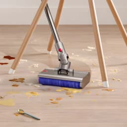 Brosse humide motorisée Dyson V12s Submarine en action sur parquet clair, rouleau bleu absorbant et aspirant liquides et miettes sous table à trois pieds