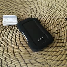 ADATA HD330 2TB noir, vue du dessus: coque en caoutchouc noir sur panier en osier avec livret de garantie en arrière-plan