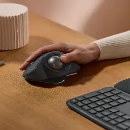 Logitech Rato Trackball MX Ergo S Grafite, uso em secretária: mão a segurar o rato junto a teclado sobre mesa de madeira