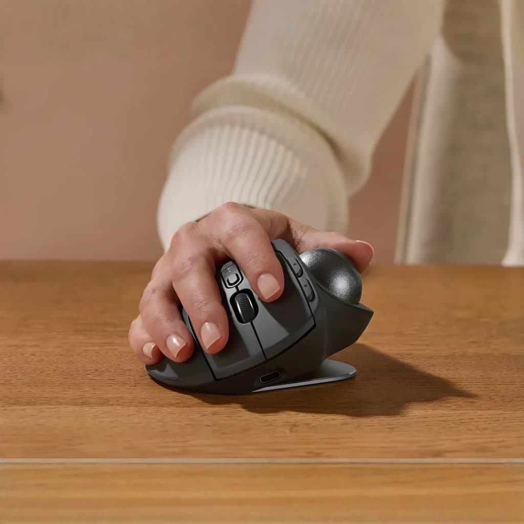 Logitech Rato Trackball MX Ergo S Grafite, uso na mão: mão segurando o rato ergonómico sobre mesa de madeira