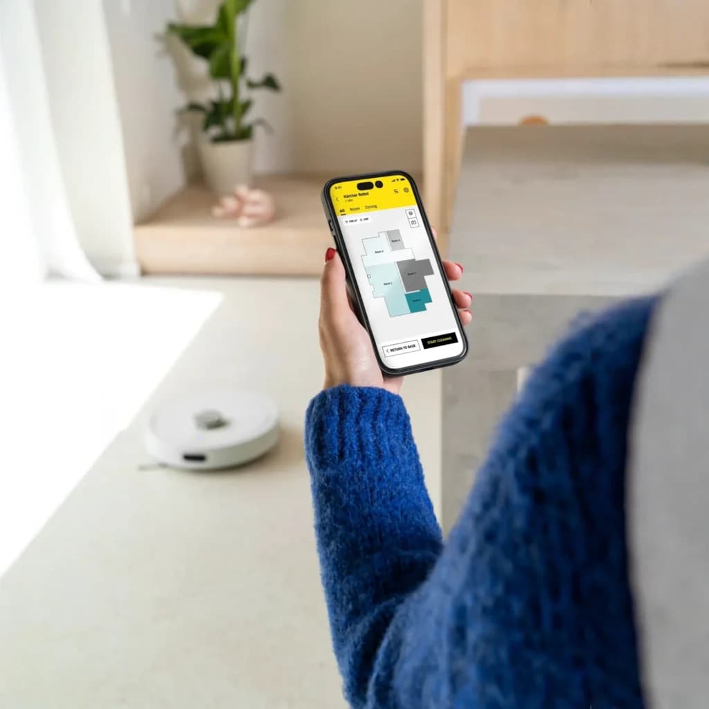 Kärcher RVC 3 Branco, uso com app: robô aspirador no chão enquanto mão segura telemóvel com mapa de divisões
