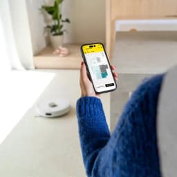Kärcher RVC 3 Branco, uso com app: robô aspirador no chão enquanto mão segura telemóvel com mapa de divisões