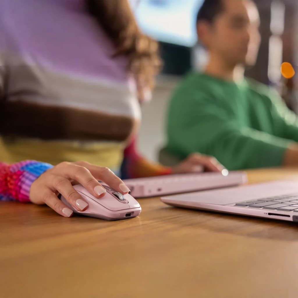 Logitech Rato MX Anywhere 3S Rosa, uso: mão segurando o rato sobre mesa de madeira ao lado de portátil rosa