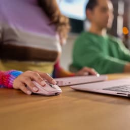 Logitech Rato MX Anywhere 3S Rosa, uso: mão segurando o rato sobre mesa de madeira ao lado de portátil rosa