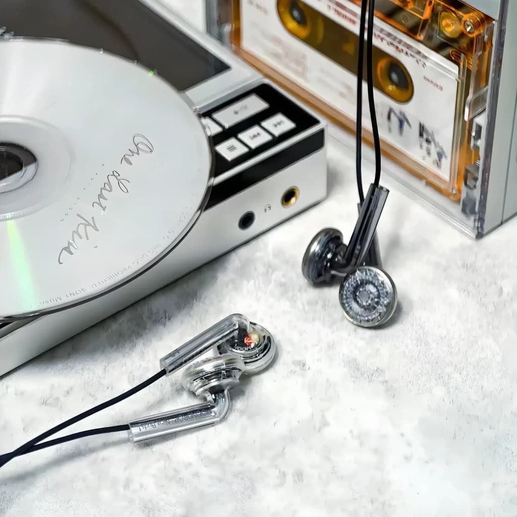 Auriculares Moondrop Nice Buds In-Ear con carcasa transparente, diseño retro tipo earbud, cable negro y conector jack 3,5 mm junto a casete y CD en escena