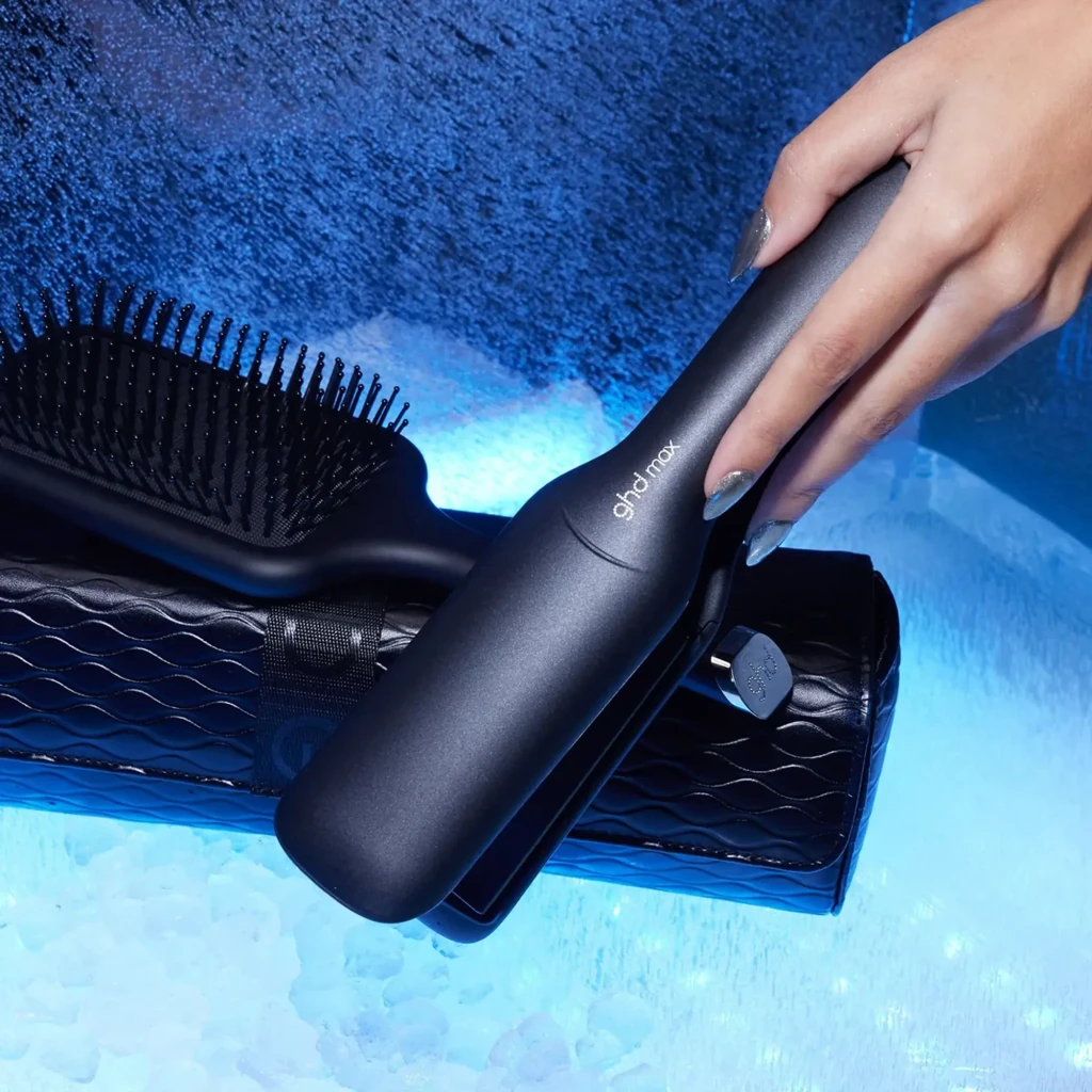GHD Max + Paddle Brush Gift Set preto, vista lifestyle: mão segurando a prancha sobre estojo e escova em fundo azul gelo