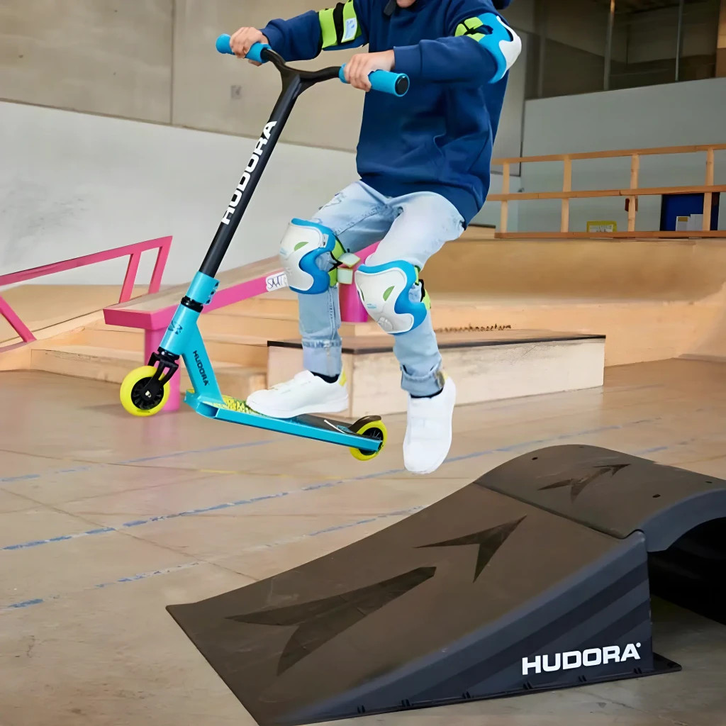 Patinete Hudora Stunt Scooter Kids 6-10 años Azul Claro, en uso: niño saltando rampa en skatepark con rodilleras
