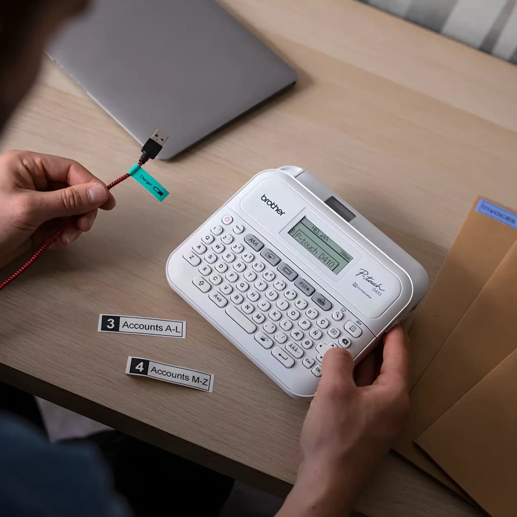 Impresora de Etiquetas Brother PT-D410VP Blanco, en uso: manos etiquetando cables y carpetas sobre escritorio con laptop