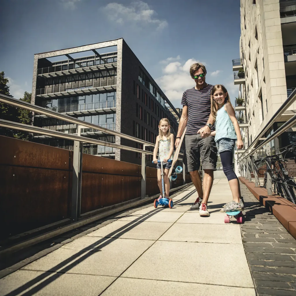 Patinete infantil Hudora Flitzkids 2.0 Azul, en uso: niña montándolo en paseo urbano junto a padre y otra niña con skateboard