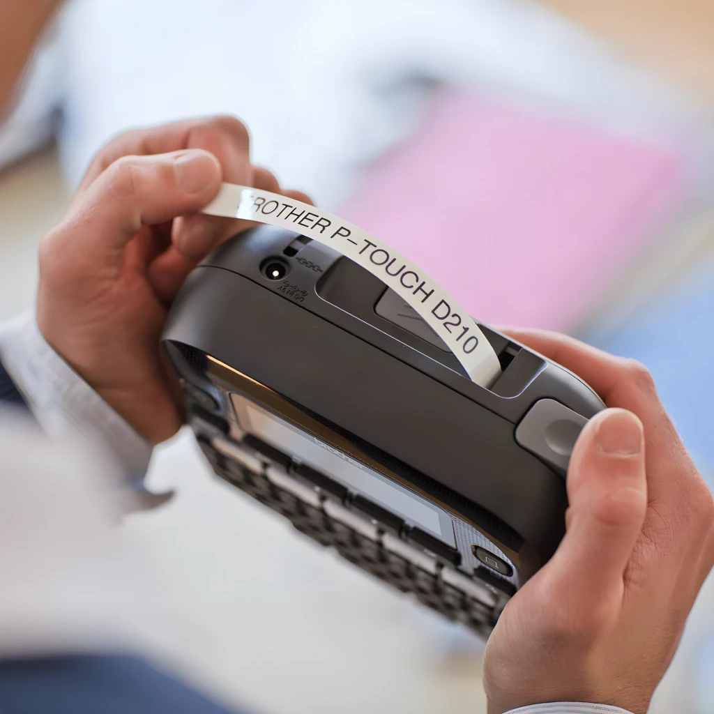 Impresora de etiquetas Brother P-touch PT-D210 Monocromo Gris/Blanco, en uso: manos sosteniendo el dispositivo mientras sale